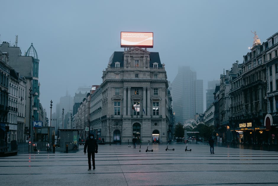 Unrecognizable pedestrians in face masks walking on city square near aged buildings against foggy sky in evening in Brussels