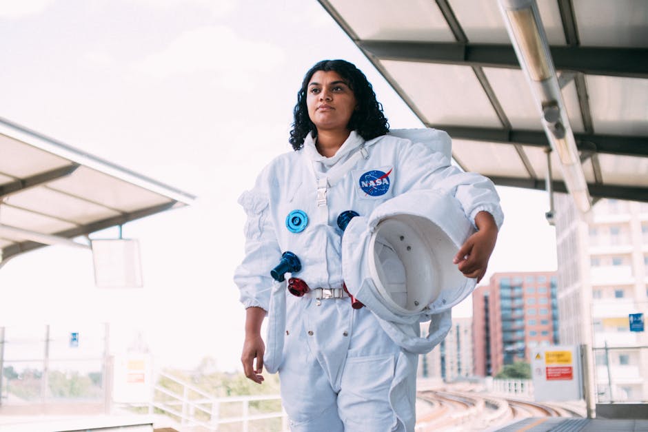A woman wearing a white astronaut suit holds a helmet at a suburban train station platform.