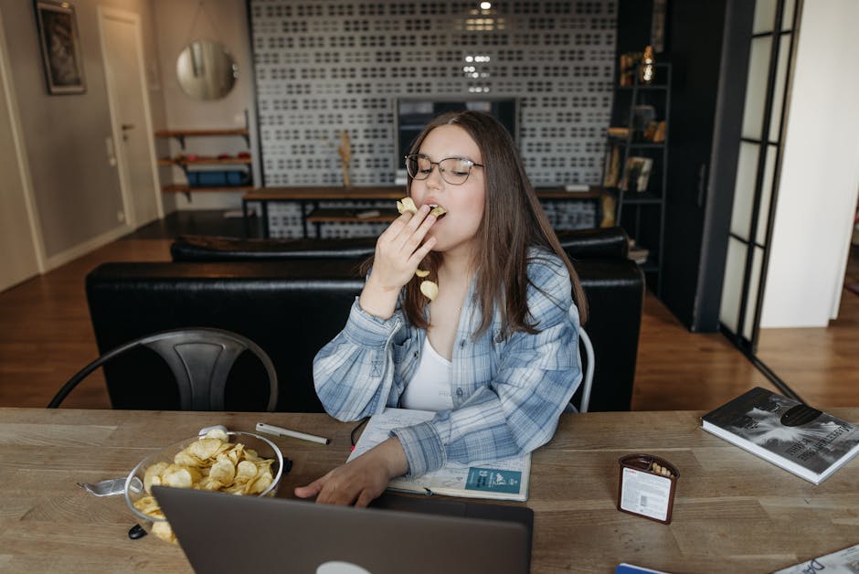 Woman in casual attire eating chips while working on a laptop indoors, suggesting a relaxed lifestyle.